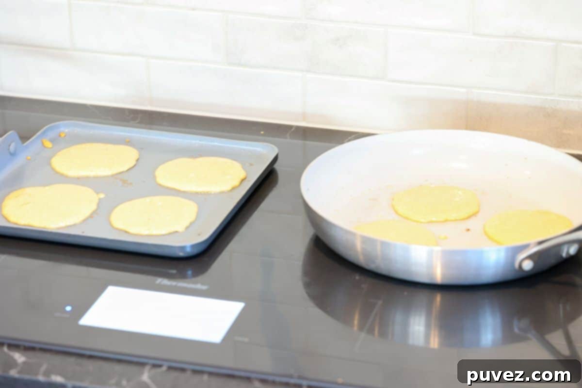 A square griddle pan and a large round skillet on a stovetop cooking pancakes.