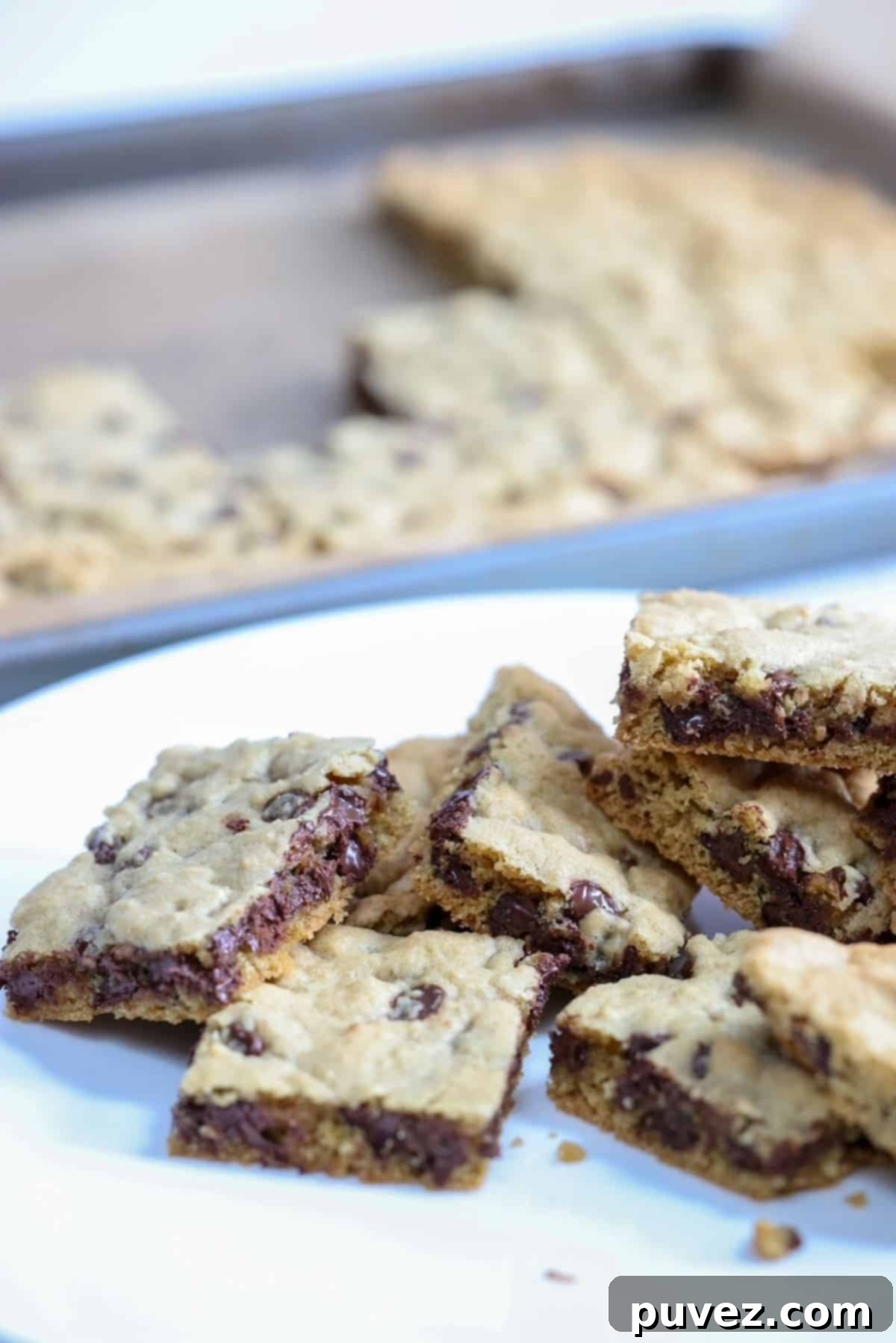 Gooey Chocolate Chip Cookie Bars 2 Chocolate Chip Cookie Bars on a white plate with a sheet pan of chocolate chip cookie bars in the background.