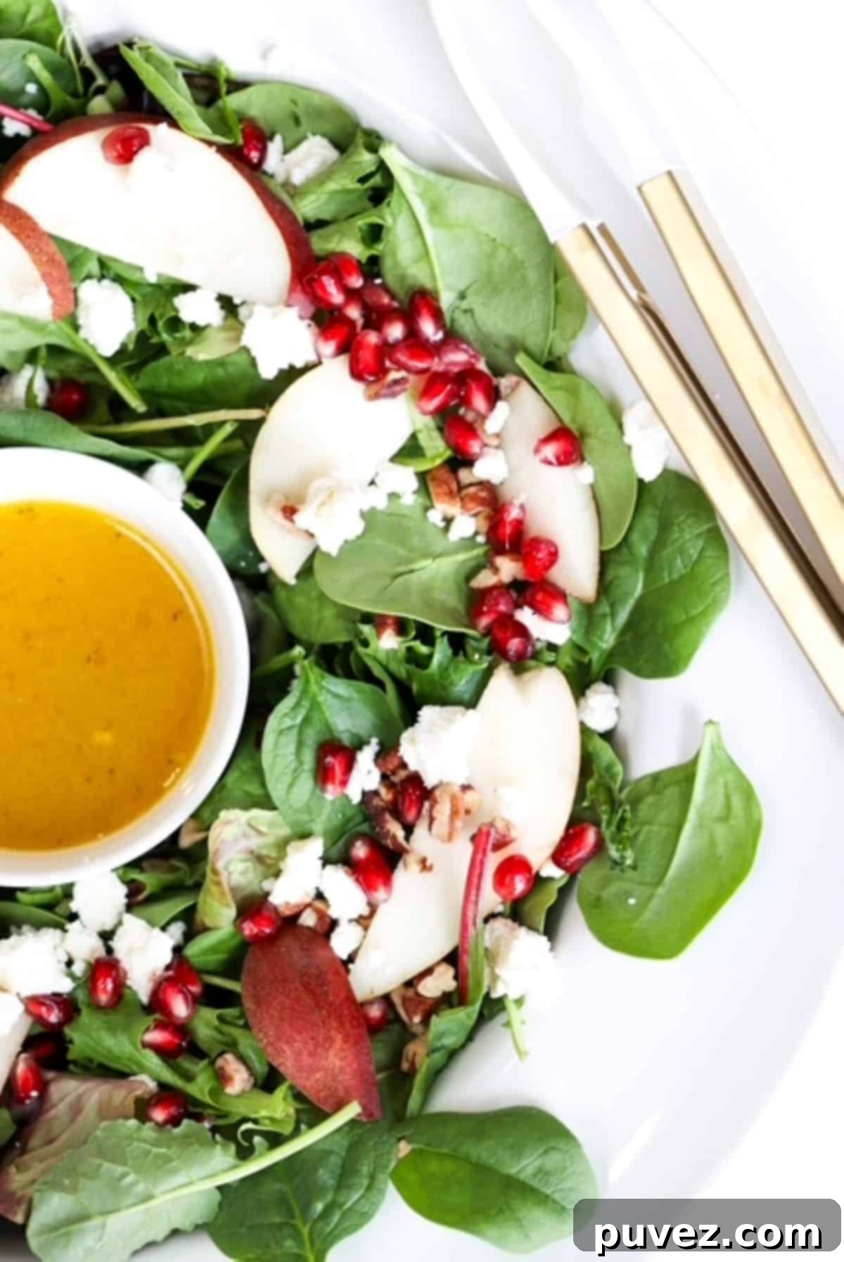 A beautifully arranged Christmas holiday salad in a white serving bowl, featuring vibrant greens, red pomegranate seeds, and white feta cheese, with gold serving tongs nearby.