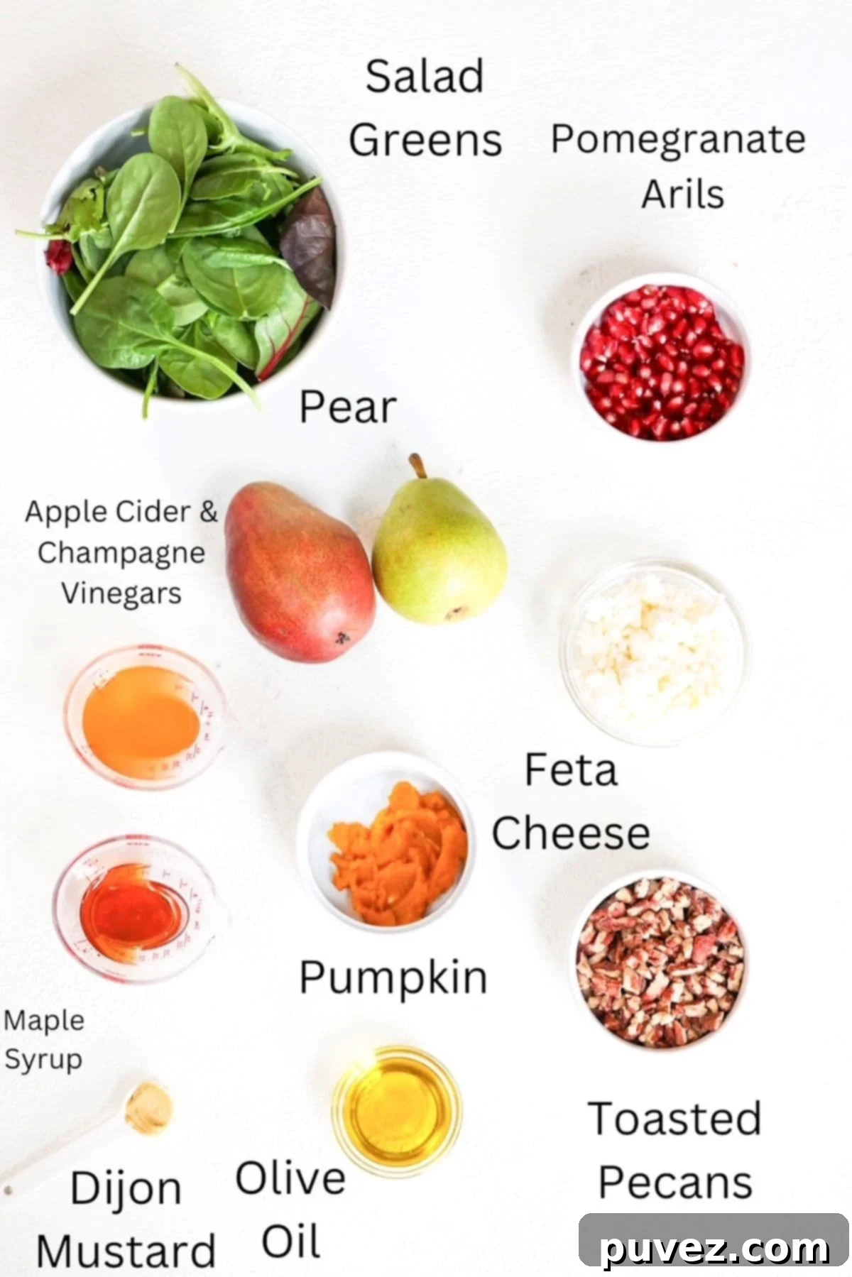 Christmas salad ingredients neatly arranged and labeled on a white background, including salad greens, a pear, pomegranate seeds, feta, pecans, and dressing ingredients.