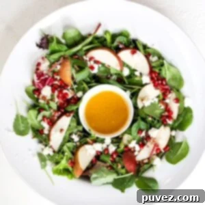 A beautifully presented Christmas salad arranged like a wreath in a large white serving bowl, with a small white bowl of dressing in the center, featuring greens, pear, pomegranate, and feta.