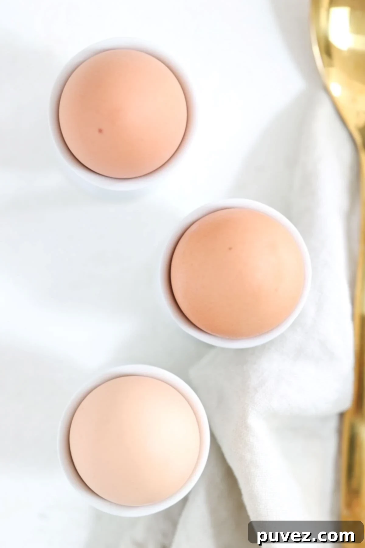 Three brown hard-boiled eggs in white egg cups with a linen napkin and gold spoon to the side.