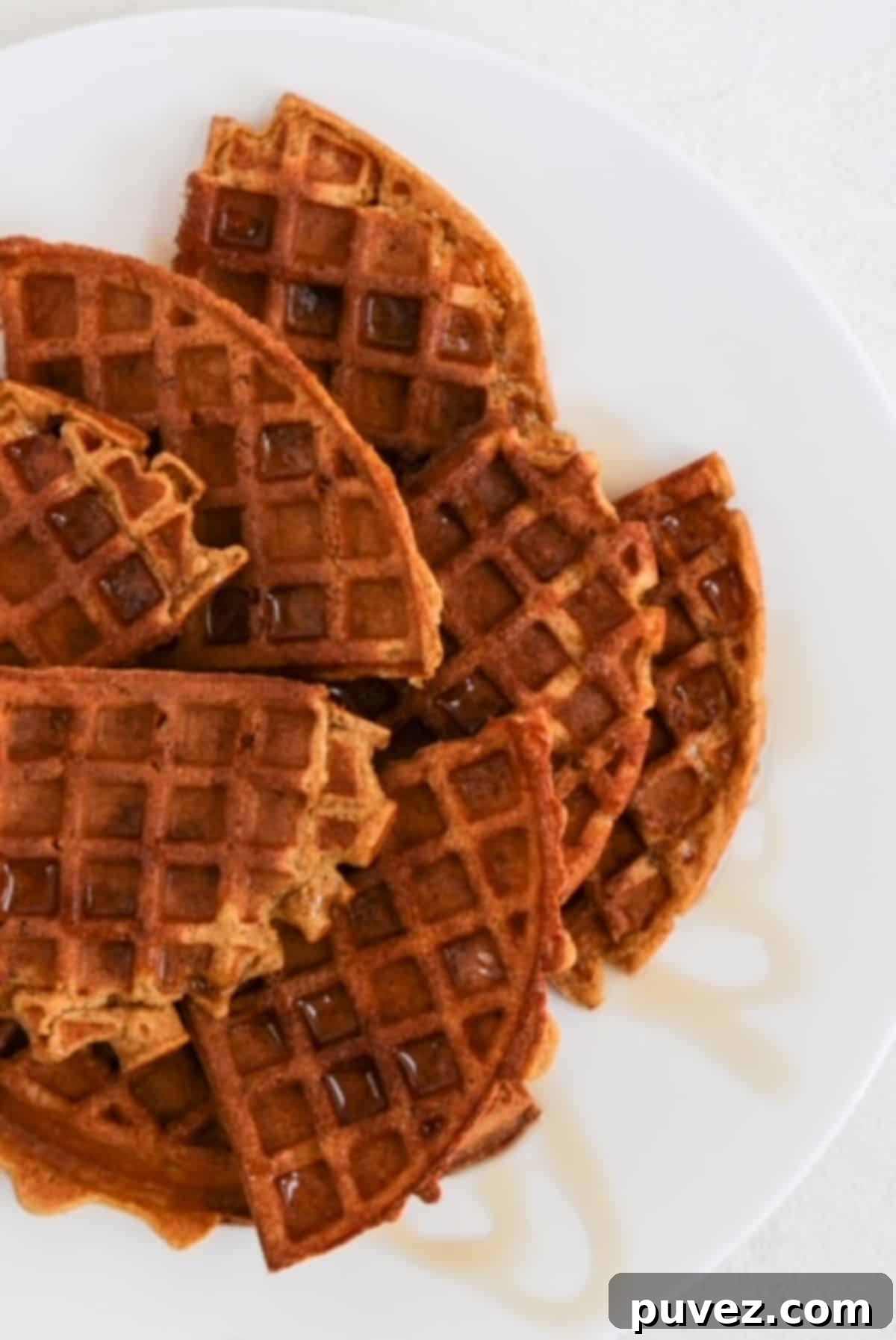 Cozy Autumn Whole Wheat Pumpkin Waffles Recipe 2 Several quarters of round pumpkin waffles overlapping on a white plate and topped with a drizzle of maple syrup.