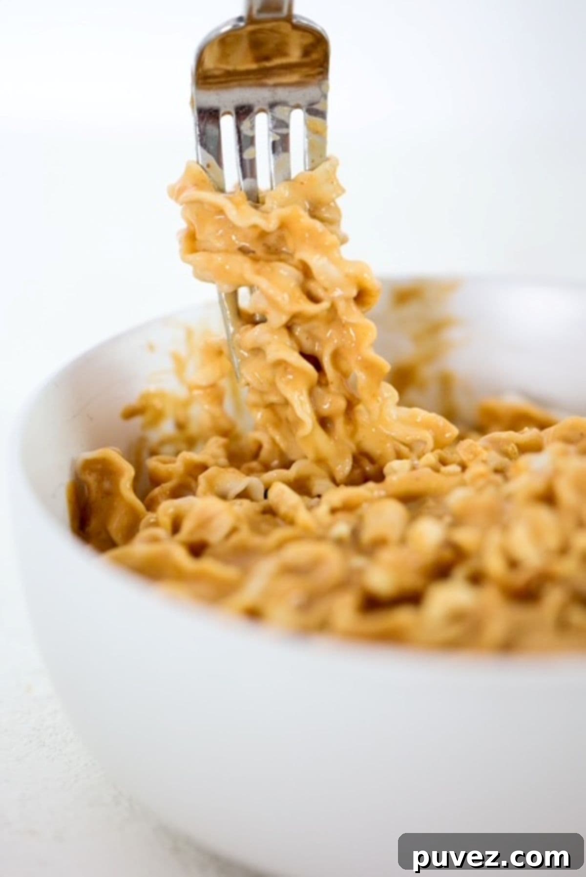 A fork with noodles wrapped around it coming out of a bowl of noodles in a peanut butter sauce.