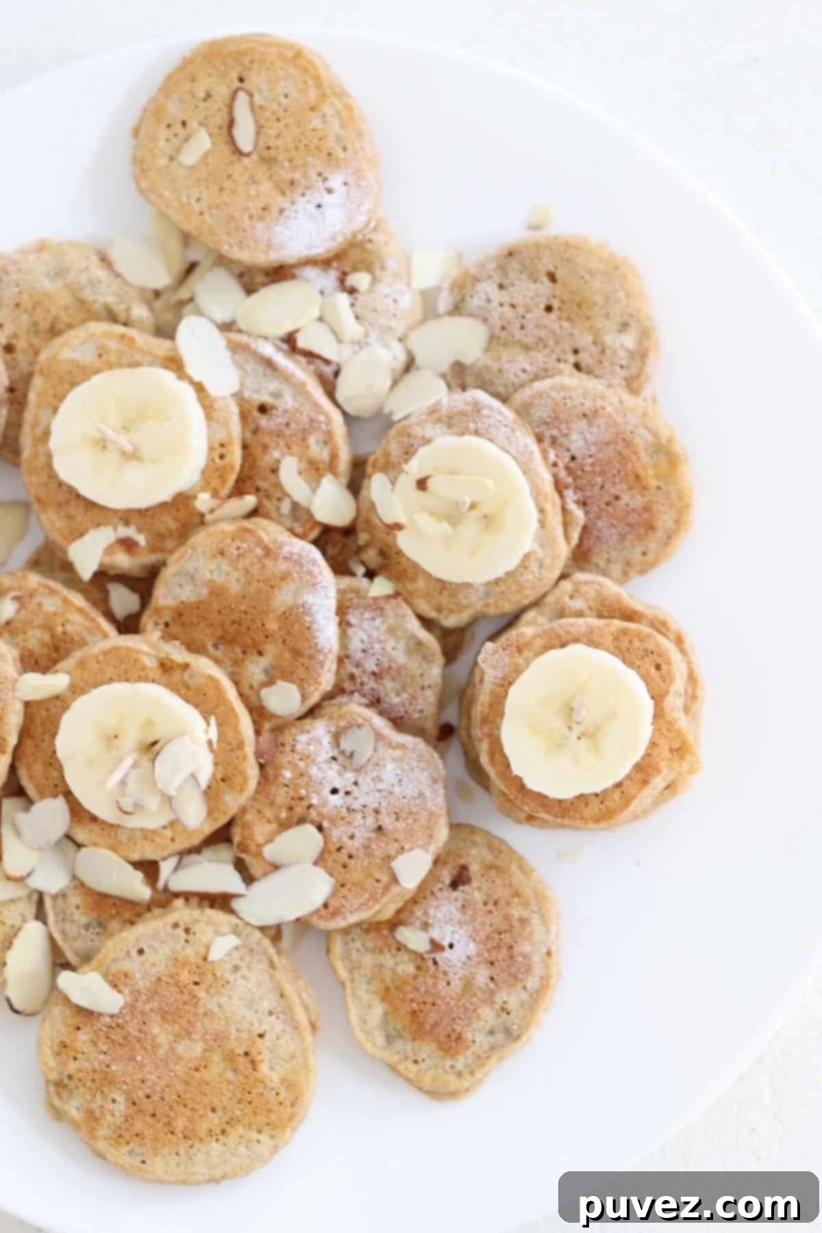 Mini banana pancakes dusted with powdered sugar, slices of banana, and sliced almond on a white plate. A perfect healthy breakfast option.