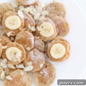 Mini banana pancakes dusted with powdered sugar, topped with sliced banana and almond slices on a white plate, highlighting a healthy and delicious breakfast.
