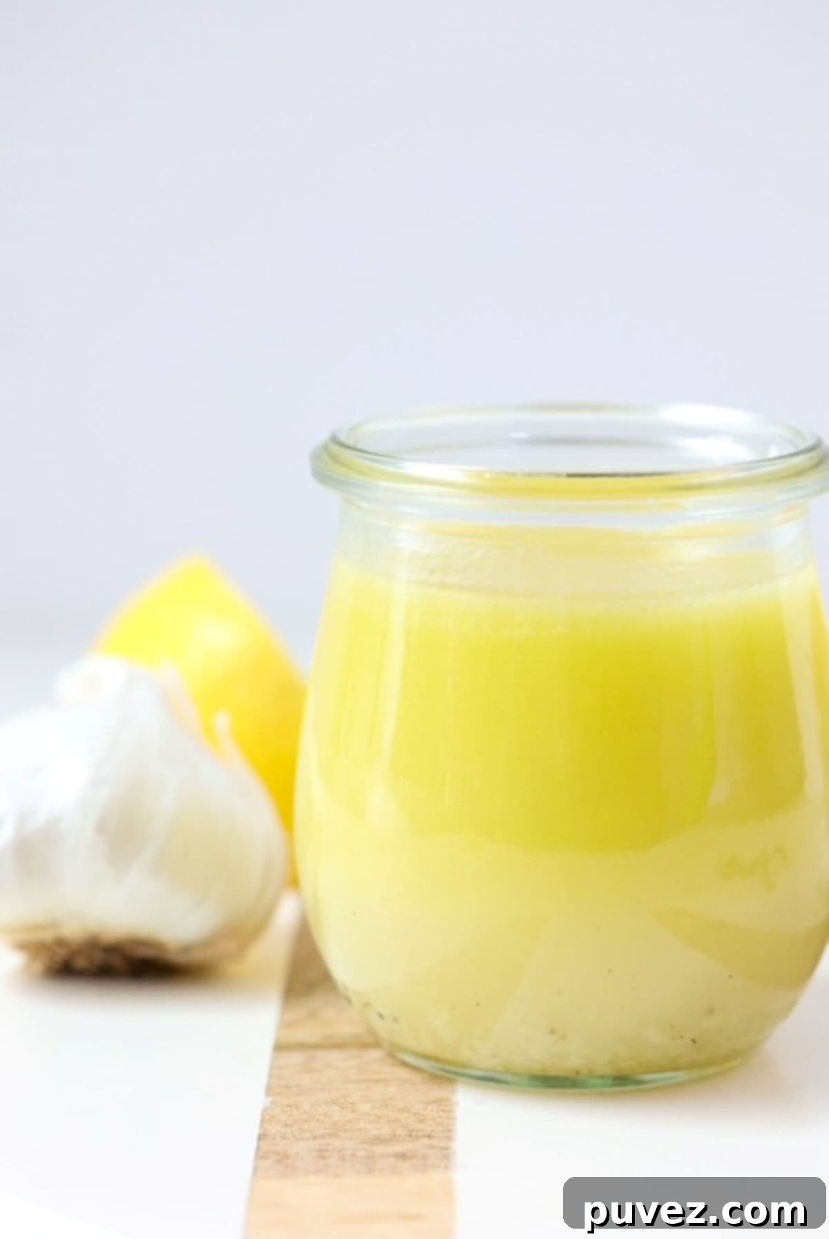 Lemon Garlic Salad Dressing on a white board with a wooden stripe. One garlic bulb and lemon sit to the back right.