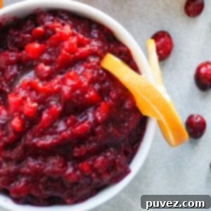 Cranberry sauce in a white bowl with one slice of orange on the right edge of the bowl. Four cranberries to the right of the bowl.