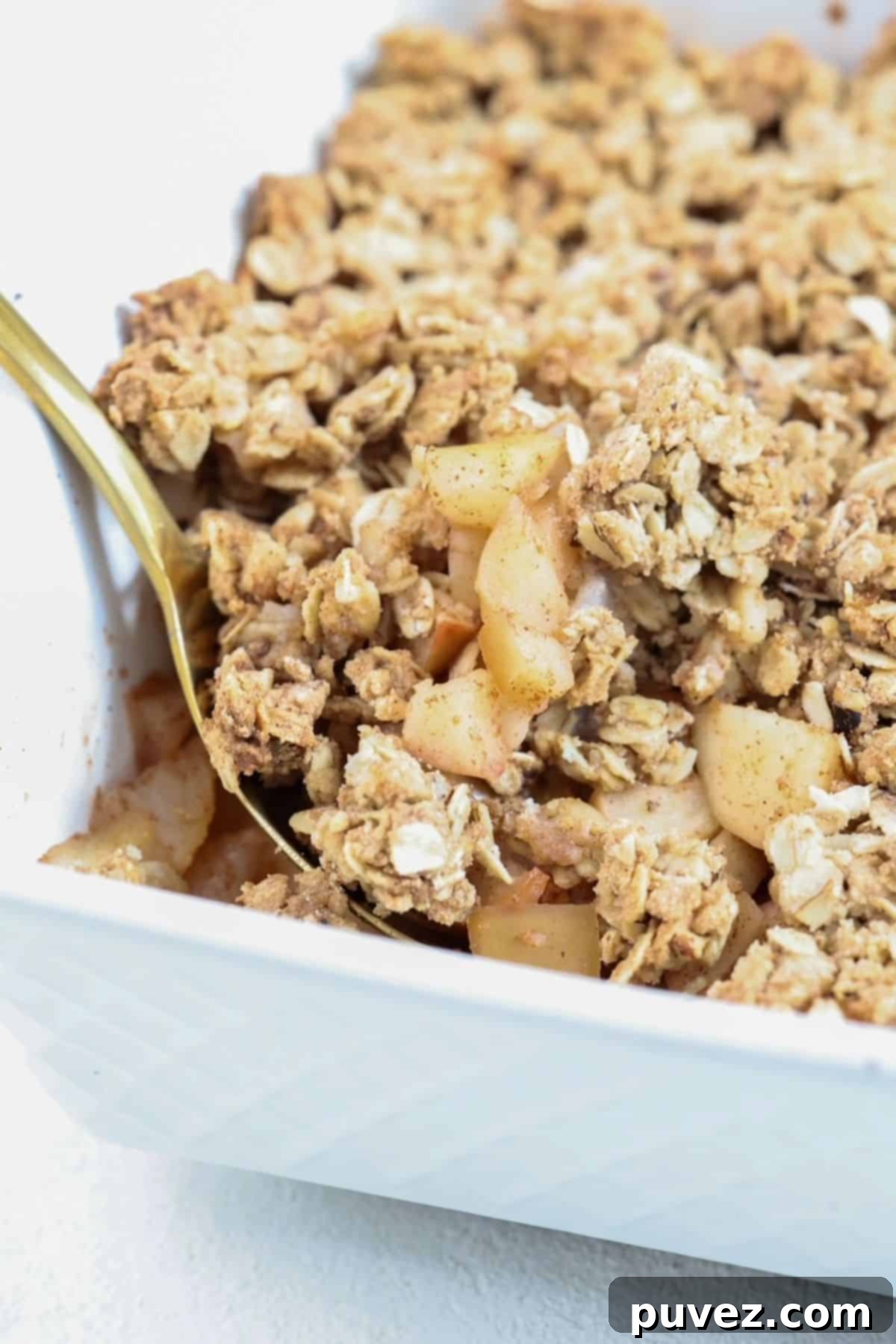 A close-up shot of a white baking dish filled with golden-brown apple and pear crumble, served with a golden spoon, highlighting the crisp oat topping and soft fruit filling.