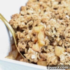 A deliciously baked apple and pear crumble, golden brown and bubbly, presented in a white baking dish with a gold spoon resting inside.