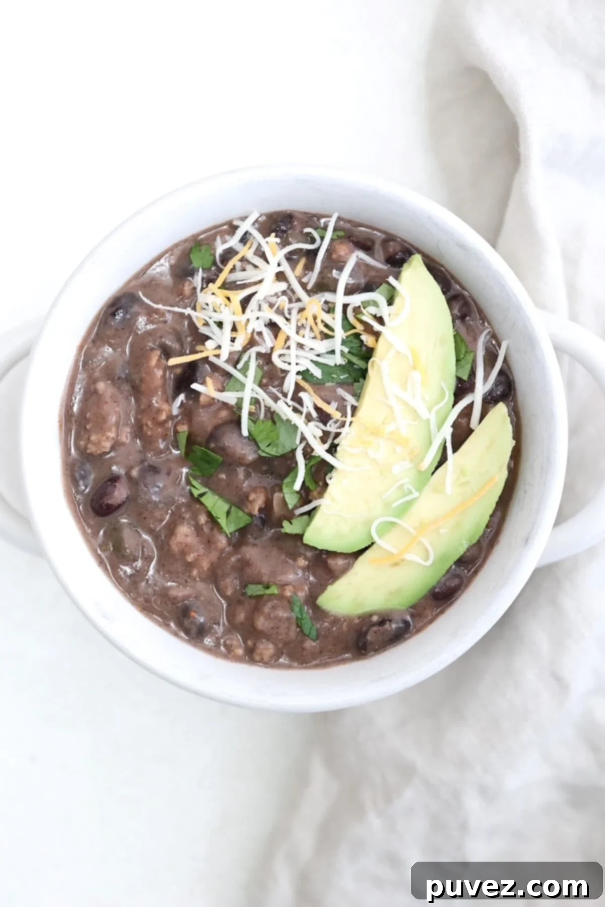 One bowl of turkey chili with black beans in a white soup bowl topped with slices of avocado and a sprinkle of cheese.